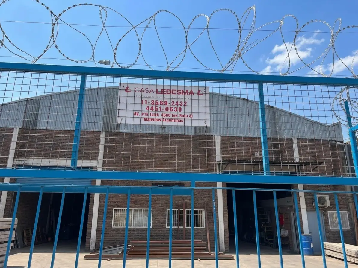 Entrada del local de Casa Ledesma: portón azul con concertina arriba y, detrás, el galpón de ladrillo con el cartel que dice Casa Ledesma, los teléfonos 11 3569-2432 y 4451-0630 y la dirección Av. Pte. Illia 4560